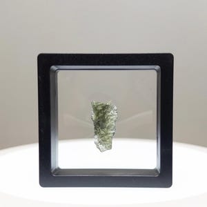 May include: A rough, olive-green gemstone is presented in a black, square, floating frame. The translucent stone has a textured surface. The frame is set against a white background.