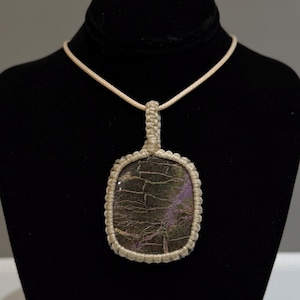 May include: A rectangular pendant necklace featuring a dark, textured stone with purple and green hues, encased in a beige macrame setting. The pendant hangs from a matching beige cord, displayed on a black mannequin.