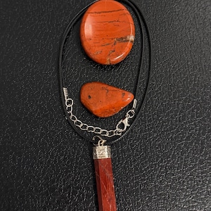 May include: A black cord necklace featuring a red jasper pendant with a silver-toned metal cap. The set includes two additional loose red jasper stones, one oval and one irregular, all displaying dark brown veining throughout the red surfaces.