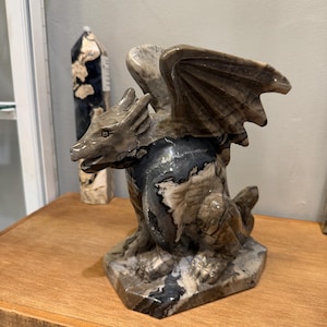 Puede incluir: Una figura de dragón tallada en piedra con una superficie pulida y patrones veteados en tonos marrón, gris, negro y blanco. La criatura está representada en posición sentada con las alas extendidas hacia arriba y una cabeza detallada.