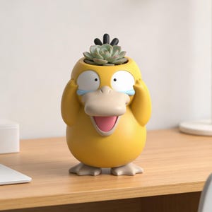 Könnte beinhalten: Ein gelber Pflanzenkübel in Form von Enton mit einer Sukkulentenpflanze auf dem Kopf. Der Kübel hat ein cartoonartiges Design mit seinen Händen, die seine Augen bedecken, und einem weit geöffneten Schnabel. Der Kübel steht auf einer Holzoberfläche.