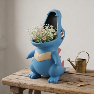 May include: A blue alligator-shaped planter with an open mouth filled with white and pink flowers. The planter has a tan belly and pink spikes. A small brass watering can sits on a wooden table next to the planter.