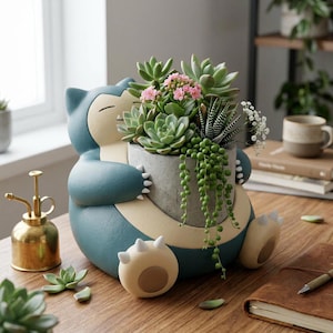 Op de afbeelding: Een blauw en crèmekleurige plantenbak in de vorm van een stripfiguur, met een grijze pot vol vetplanten en hangplanten. De plantenbak heeft een lachend gezicht en staat op een houten tafel. Links staat een gouden gieter.