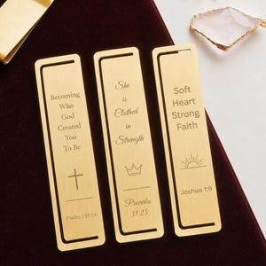 May include: Three rectangular gold-toned metal bookmarks with engraved text and symbols. The first reads "Becoming Who God Created You To Be" with a cross, the second "She Is Clothed in Strength" with a crown, and the third "Soft Heart Strong Faith" with a sunrise.