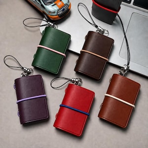 May include: A collection of six small rectangular leather cases in green, dark brown, purple, red, and light brown, each secured with a colored elastic band and featuring a black cord loop with a silver metal attachment for hanging.