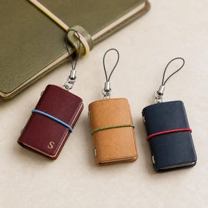 May include: Three miniature leather-bound notebooks with metal jump rings and black cord loops. The burgundy notebook features a gold letter S, a blue elastic band, and a tan notebook. The tan notebook has a green elastic band. The navy notebook has a red elastic band.