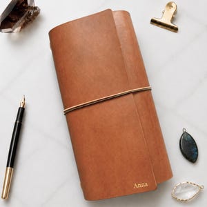 May include: A tan leather journal cover with a vertical elastic band closure, featuring the name Anna embossed in gold on the bottom right corner. The journal is accompanied by a black and gold fountain pen, a gold metal clip, and two crystals.