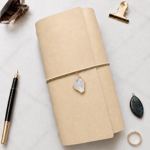 May include: A beige leather journal cover featuring a vertical elastic band closure with a dangling clear crystal charm. The cover is embossed with the name Anna in the bottom right corner. The design includes a smooth, matte finish.