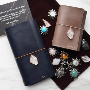 May include: Two leather journals, one navy blue and one brown, with crystal pendants and elastic closures. The journals are personalized with names. Various gemstone sunburst charms and loose crystals are scattered around the journals.