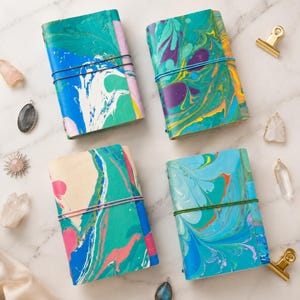 May include: Four rectangular journals featuring marbled covers in vibrant shades of blue, green, purple, pink, and yellow. Each journal is secured with a thin elastic band in black, blue, pink, or green, and surrounded by various crystal pendants.