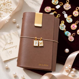 May include: A brown leather journal featuring a gold metal clip, a thin elastic band holding two rectangular white and gold charms with the letters J and M, and the date 29.5.2024 embossed in gold. A card with Happy Anniversary text is included.