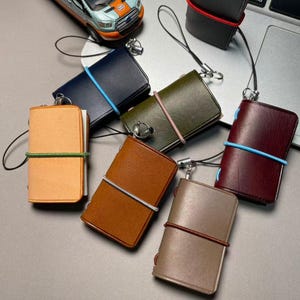 Op de afbeelding: Een verzameling van zeven kleine notitieboekjes met een leren kaft in diverse kleuren, waaronder beige, marineblauw, olijfgroen, bruin, bordeauxrood en grijs, elk vastgezet met een gekleurde elastieken band en voorzien van een metalen bedel aan een koord.