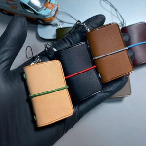 May include: Three miniature rectangular leather notebook charms with metal eyelets and elastic bands. One is light tan with a green band, one is black with a red band, and one is brown with a blue band. Each includes a small metal attachment loop.
