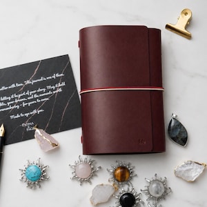 Könnte beinhalten: Ein dunkelburgunderfarbenes Lederjournal, das mit einem zweifarbigen rot-weißen Gummiband verschlossen ist, begleitet von einer schwarzen Karte mit weißem Text, einem goldenen Metallclip und verschiedenen Edelsteinanhängern wie Türkis und Quarz.