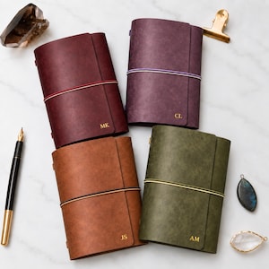 May include: Four leather journals in burgundy, purple, brown, and olive green, each secured with a colored elastic band. The covers feature gold-embossed initials MK, CL, JS, and AM. A fountain pen, a gold clip, and two crystals are arranged nearby.