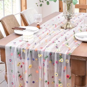 May include: A sheer white table runner with colorful embroidered flowers in shades of yellow, pink, blue, and orange. The runner is draped over a wooden table set for a meal, with plates, silverware, and glasses.