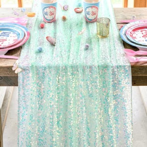 Op de afbeelding: Een tafelsetting met een mintgroene pailletten tafelloper, borden, bekers en roze bestek. De bekers hebben de tekst "He She?" erop. Decoratieve items zijn kleine vlinders en pastelkleurige bollen.
