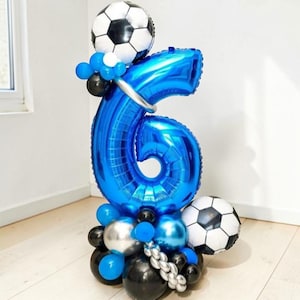 May include: A blue number 6 balloon arrangement with soccer ball accents. The arrangement includes a large blue number 6 balloon, soccer ball balloons, and smaller blue, black, and silver balloons. A festive decoration for a birthday party.