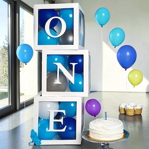 Op de afbeelding: Witte dozen met de tekst "ONE" met blauwe, grijze en witte ballonnen erin. Verschillende blauwe, turquoise en gele ballonnen zweven in de buurt. Een witte taart met een enkele brandende kaars staat op een taartstandaard, met cupcakes op de achtergrond. Feestversieringen.