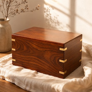 May include: A rectangular wooden box with a rich dark brown grain pattern, featuring smooth surfaces and metallic gold-colored corner accents that secure the joints on the front and side panels, creating a clean and structured appearance.