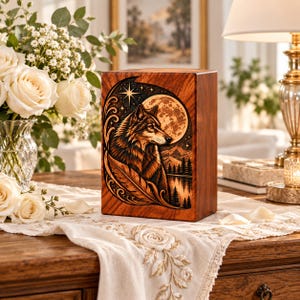 May include: A rectangular wooden cremation urn featuring a detailed laser-engraved illustration of a wolf, a full moon, a star, and a mountain landscape with pine trees. The design is framed by a large feather and decorative swirling patterns.