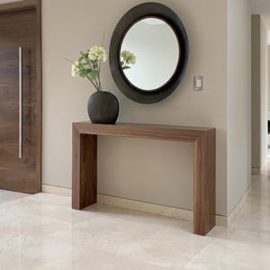 Puede incluir: Una entrada moderna con una consola rectangular de madera oscura. Encima, un espejo redondo con marco oscuro. Un jarrón oscuro con flores blancas descansa sobre la mesa, creando una estética minimalista.