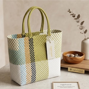 May include: A woven market tote bag with a patchwork design in shades of green, white, blue, tan, and black. The bag has braided green handles and a tag that reads "JogjaFound". The bag is sitting on a table.