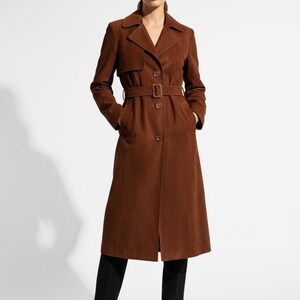 May include: A long, brown suede trench coat with a belt and button closure. The coat features a notched collar and pockets. The model is wearing black pants and black boots. The coat is a classic outerwear piece.