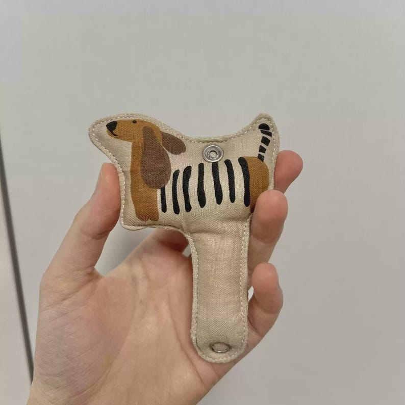 May include: A beige fabric dog-shaped keyring with brown ears and a black striped body. The keyring has a metal snap closure and a loop at the bottom. The dog design is stitched onto the fabric.