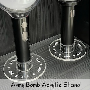 May include: Two black cylindrical light sticks with white spherical tops, each mounted on a circular clear acrylic stand. The stands feature the white printed text "BTS" and "ARMY" along with small logo icons arranged in a repeating circular pattern.
