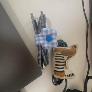 Puede incluir: Dos organizadores de cables: una flor de cuadros azules y blancos y una bota de fieltro con detalles marrones y beige. Los organizadores sujetan cables negros y grises estampados, probablemente para electr&oacute;nica.