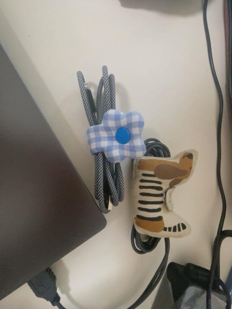 Puede incluir: Dos organizadores de cables: una flor de cuadros azules y blancos y una bota de fieltro con detalles marrones y beige. Los organizadores sujetan cables negros y grises estampados, probablemente para electr&oacute;nica.