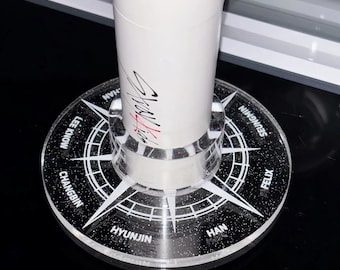 Könnte beinhalten: Ein runder, schwarzer Acrylständer mit Glitzer-Finish, der ein weißes Kompass-Design und die Namen Bang Chan, Lee Know, Changbin, Hyunjin, Han, Felix und Seungmin am Rand aufweist und einen weißen Leuchtstab trägt.