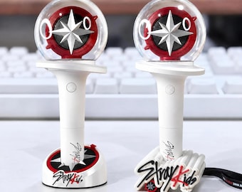 Könnte beinhalten: Zwei weiße Stray Kids Leuchtstäbe mit einer transparenten, kugelförmigen Spitze, die einen rot-silbernen Kompassstern enthält. Die weißen Griffe haben einen runden Knopf und das Stray Kids Logo in Schwarz und Rot aufgedruckt.
