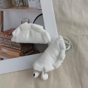 Op de afbeelding: Een wit pluche hoesje in de vorm van een dumpling voor draadloze oordopjes. De zachte, pluizige hoes heeft een ontwerp met geplooide randen en wordt getoond met een paar witte draadloze oordopjes en een oplaadkabel die uit het hoesje steekt.
