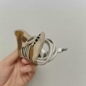 May include: A beige fabric cable tidy with a dog-like design, featuring brown and black accents. It holds a white charging cable, offering a neat and stylish way to manage cords.