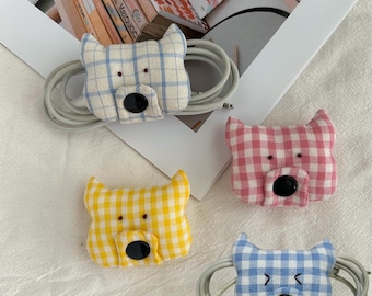 Könnte beinhalten: Vier Kabel-Organizer aus Stoff in Form von Katzenköpfen mit Vichy-Karomustern in Blau, Pink, Gelb und Hellblau. Jeder Organizer hat eine schwarze Knopf-Nase, gestickte Augen und spitze Ohren, um Ladekabel ordentlich aufzuwickeln.