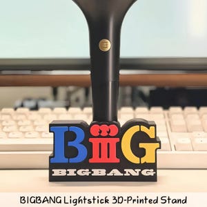 May include: A black 3D-printed stand for a lightstick featuring the word BIG in large, colorful letters with blue for B, red for I, and yellow for G. The base displays the text BIGBANG in white capital letters on a black background.