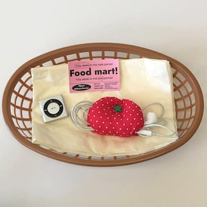Könnte beinhalten: Ein ovaler brauner Kunststoffkorb mit einem cremefarbenen Papierbogen, einem silbernen quadratischen digitalen Musikplayer, einem rosa Food mart! Flyer und einem roten, tomatenförmigen Kabel-Organizer aus Stoff mit weißen Punkten.