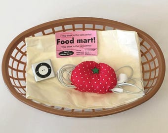 May include: A brown oval plastic basket containing a cream-coloured paper sheet, a silver square digital music player, a pink Food mart! flyer, and a red fabric tomato-shaped cord organiser with white polka dots and a green star button.