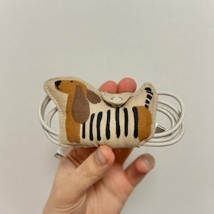 May include: A beige fabric cable tidy in the shape of a dachshund dog, featuring brown ears and a black striped body. It holds a white cable with a USB connector.