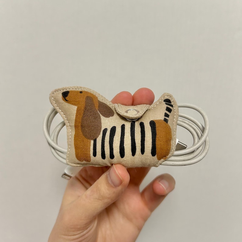 May include: A beige fabric cable tidy in the shape of a dachshund dog, featuring brown ears and a black striped body. It holds a white cable with a USB connector.