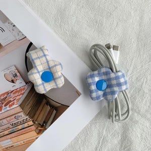Puede incluir: Dos organizadores de cables con forma de flor. Cada organizador est&aacute; hecho de tela a cuadros azul y blanco con un bot&oacute;n azul en el centro. Uno sostiene un cable gris con conectores USB blancos. El otro organizador est&aacute; vac&iacute;o.