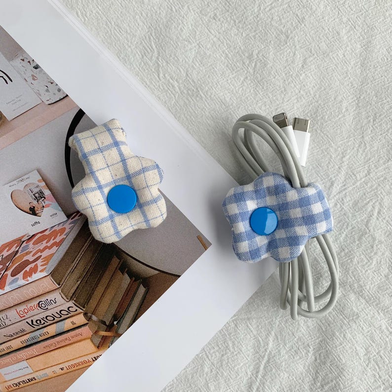 Puede incluir: Dos organizadores de cables con forma de flor. Cada organizador est&aacute; hecho de tela a cuadros azul y blanco con un bot&oacute;n azul en el centro. Uno sostiene un cable gris con conectores USB blancos. El otro organizador est&aacute; vac&iacute;o.