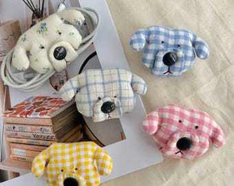 May include: A selection of dog-shaped fabric cable tidies in various colours and patterns. The tidies feature a button nose and embroidered details. The patterns include floral, plaid, and gingham designs. The colours are white, blue, yellow, and pink.