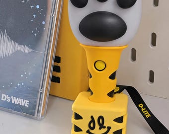 Könnte beinhalten: Ein gelb-schwarzer Leuchtstab mit einem weißen Katzenpfoten-Aufsatz und schwarzen Ballen. Die Basis ist ein gelbes Quadrat mit einem schwarzen Smiley-Logo und dem Schriftzug D-LITE auf einer schwarzen Handschlaufe. Der Griff hat einen gelben Knopf.