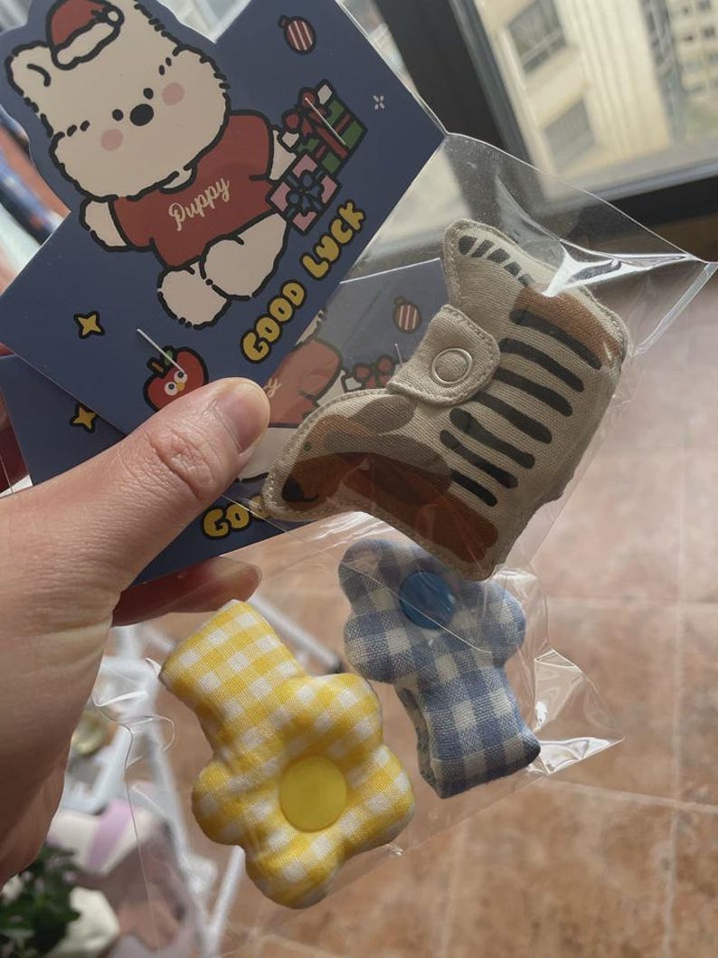 Puede incluir: Una bolsa de pl&aacute;stico transparente contiene un zapato a rayas beige y marr&oacute;n, una flor de cuadros amarillos y una flor de cuadros azules y blancos. Tambi&eacute;n se ve una tarjeta azul con un cachorro de dibujos animados con un gorro de Pap&aacute; Noel y las palabras "Good Luck".