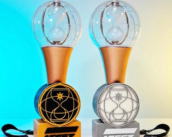 Könnte beinhalten: Zwei ATEEZ-Lightsticks mit klaren Glaskuppeln, die Sanduhren enthalten, montiert auf 3D-gedruckten Ständern. Ein Ständer ist goldfarben mit schwarzem Emblem, der andere silberfarben mit weißem Emblem. Beide tragen den Schriftzug ATEEZ.