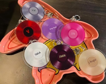 May include: A collection of seven circular acrylic keychains shaped like vinyl records, each featuring a different color and the name RM, V, Jung Kook, JIN, SUGA, JIMIN, or j-hope, resting on a pink, star-shaped ceramic tray with a yellow center.