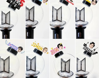 Könnte beinhalten: Eine Sammlung von acht BTS-Leuchtstäben, jeder mit einer klaren, kugelförmigen Spitze, einem schwarzen BTS-Logo im Inneren und einem schwarzen Griff. Jeder Leuchtstab ist mit einem individuellen Acryl-Namensschild und einem Foto eines Mitglieds versehen: BTS, RM, V, JK, J-Hope, Jimin, Jin und Suga.
